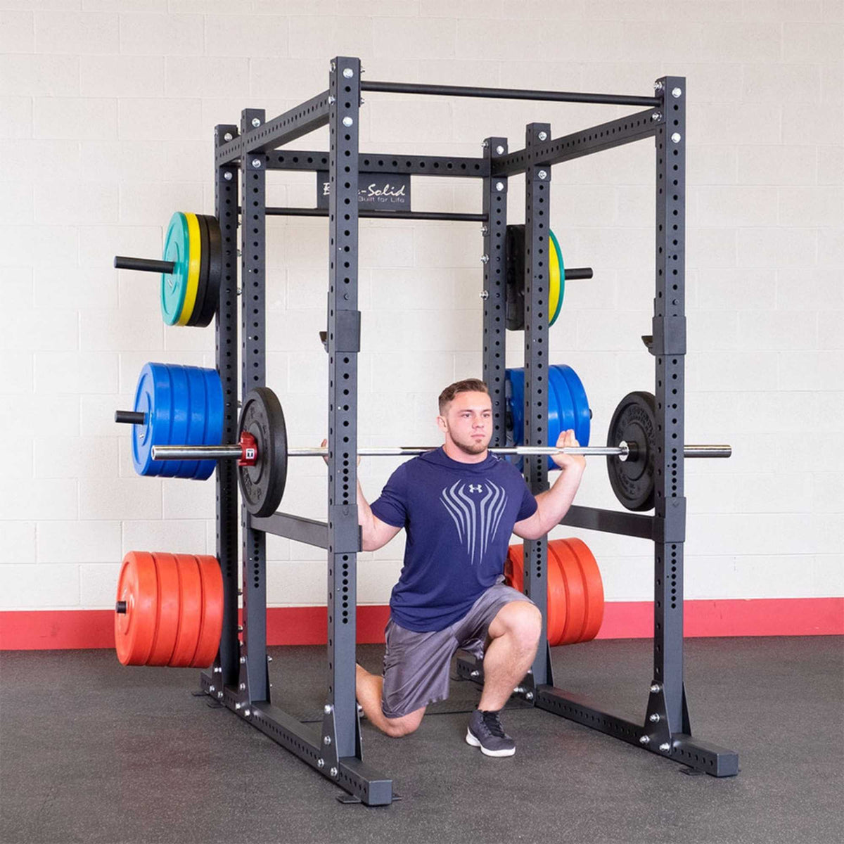 Body-Solid Pro ClubLine SPRBACK Power Rack Rear Extension SPRBACK Man performing a barbell squat inside a Body-Solid power rack loaded with colorful weight plates in a gym setting.
