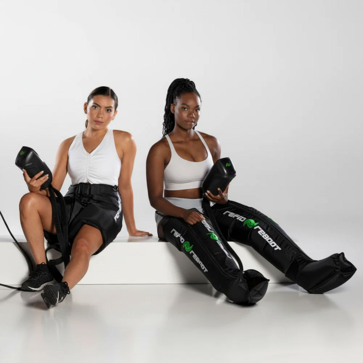 Two women sitting on a white floor with black leg compression boots and hip attachment holding black control devices.