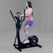 Best Fitness Cross Trainer Elliptical BFCT1B Three-quarter view of woman training on Best Fitness CT1 elliptical trainer in a gray gym setting.

