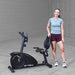 Best Fitness Recumbent Bike BFRB1B Full-body image of a woman posing next to the Best Fitness RB1 recumbent exercise bike in a minimalist studio setting.