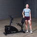 Best Fitness Recumbent Bike BFRB1B Woman standing beside the Best Fitness RB1 recumbent exercise bike, smiling after a workout session.