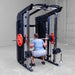 A man performing a squat on a Body-Solid-GSFR-100All-in-OneFunctional-Trainer-Smith-Machine with red weight plates in a garage setting.