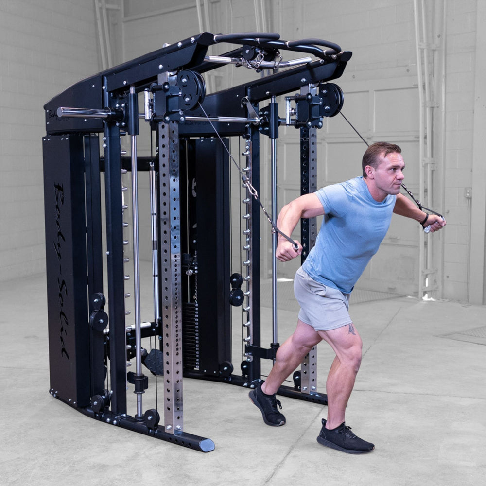 A man in a light blue t-shirt and gray shorts exercises with a Body-Solid-GSFR-100All-in-OneFunctional-Trainer-Smith-Machine in a gym setting. The cable machine is black and silver with the brand name 'Endur' visible on the side.