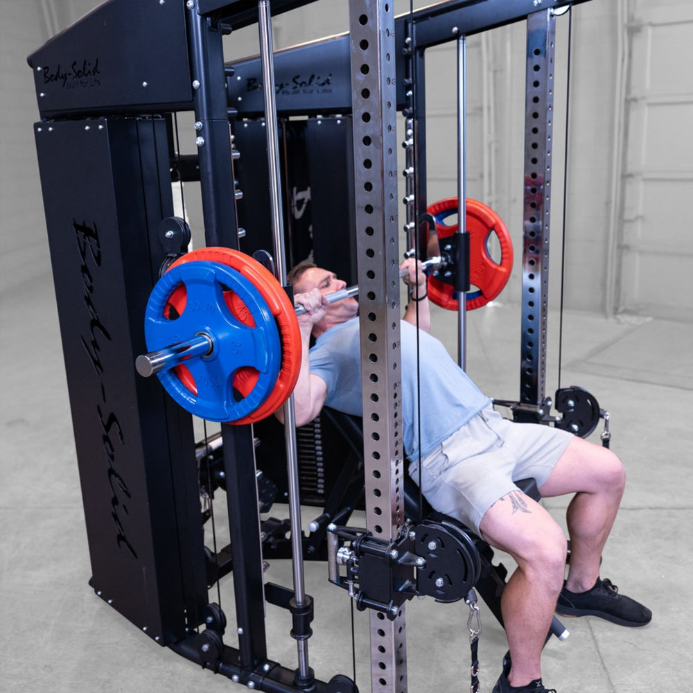 A man performing a bench press on a Body-Solid-GSFR-100All-in-OneFunctional-Trainer-Smith-Machine with red and blue weight plates in a gym setting.