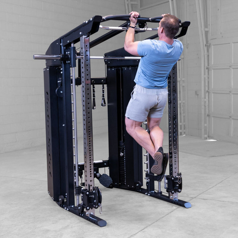 A man in a blue t-shirt and gray shorts performs a pull-up on a Body-Solid-GSFR-100All-in-OneFunctional-Trainer-Smith-Machine inside a garage.