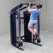 A man in a blue t-shirt and gray shorts performs a pull-up on a Body-Solid-GSFR-100All-in-OneFunctional-Trainer-Smith-Machine inside a garage.