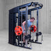 A man in a light blue t-shirt and gray shorts performs a cable push-up on a Body-Solid-GSFR-100All-in-OneFunctional-Trainer-Smith-Machine with red weight plates, set in a warehouse gym environment.