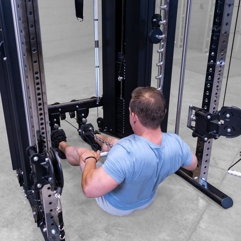 A man in a light blue t-shirt sits on the floor using a cable pulley on a Body-Solid-GSFR-100All-in-OneFunctional-Trainer-Smith-Machine in a gym setting.