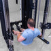 A man in a light blue t-shirt sits on the floor using a cable pulley on a Body-Solid-GSFR-100All-in-OneFunctional-Trainer-Smith-Machine in a gym setting.
