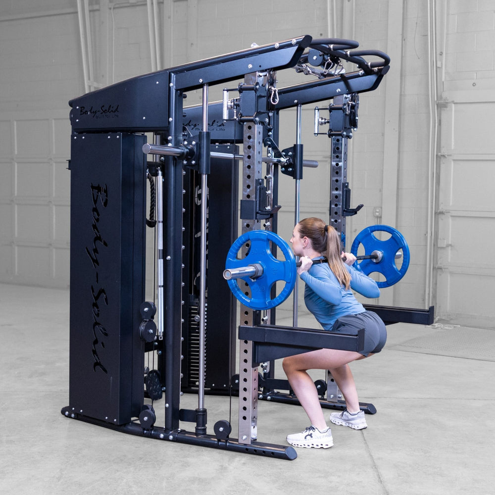 A woman performing a squat on a barbell rack attachment within Body-Solid-GSFR-100All-in-OneFunctional-Trainer-Smith-Machine set in a concrete-floored warehouse gym environment.