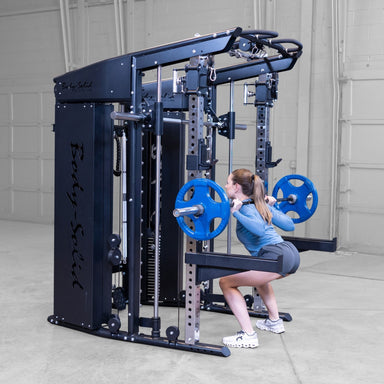 A woman performing a squat on a barbell rack attachment within Body-Solid-GSFR-100All-in-OneFunctional-Trainer-Smith-Machine set in a concrete-floored warehouse gym environment.
