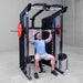 A man performing a barbell squat on a Body-Solid-GSFR-100All-in-OneFunctional-Trainer-Smith-Machine in a concrete-floored indoor gym space.