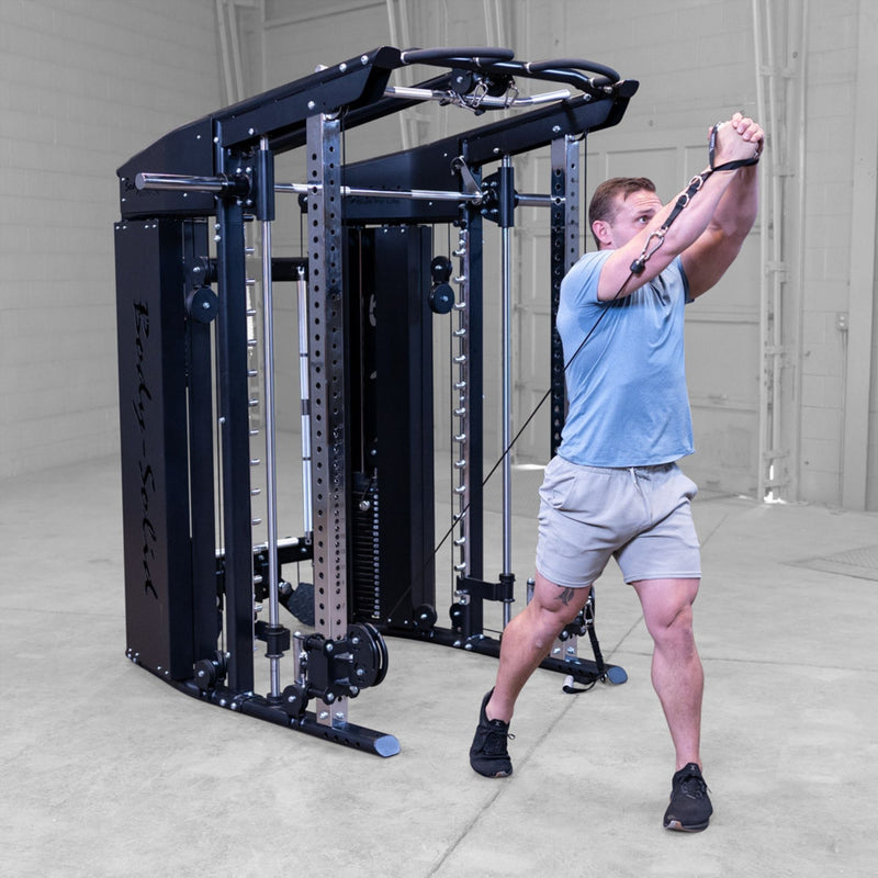 A man in a gym using a dual cable pulley system attached to a Body-Solid-GSFR-100All-in-OneFunctional-Trainer-Smith-Machine, wearing a light blue t-shirt, gray shorts, and black sneakers.