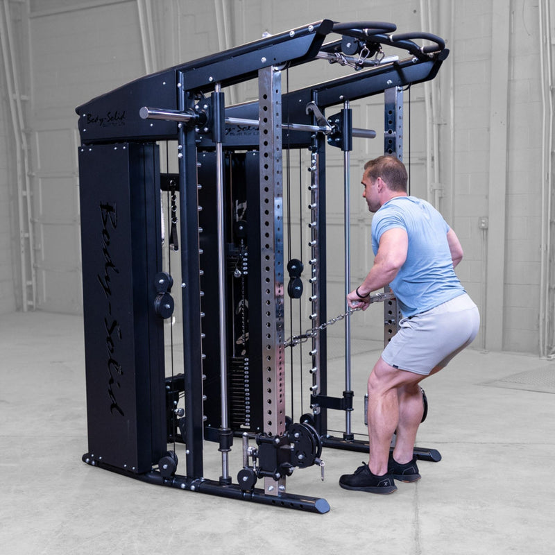 A man in a light blue t-shirt and gray shorts performing a cable pull-through exercise on a Body-Solid-GSFR-100All-in-OneFunctional-Trainer-Smith-Machine in a warehouse setting.