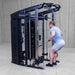 A man in a light blue t-shirt and gray shorts performing a cable pull-through exercise on a Body-Solid-GSFR-100All-in-OneFunctional-Trainer-Smith-Machine in a warehouse setting.