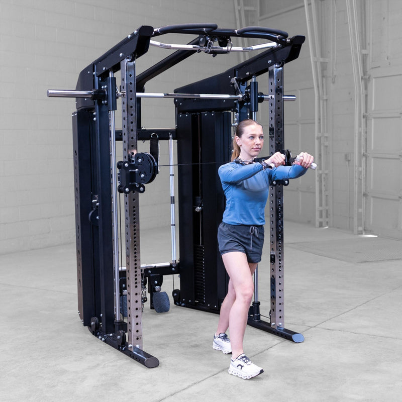 A woman in athletic wear is using a black and silver Body-Solid GSFR100 multi-functional home gym cable machine with a pull-up bar, set in a concrete-floored warehouse gym.