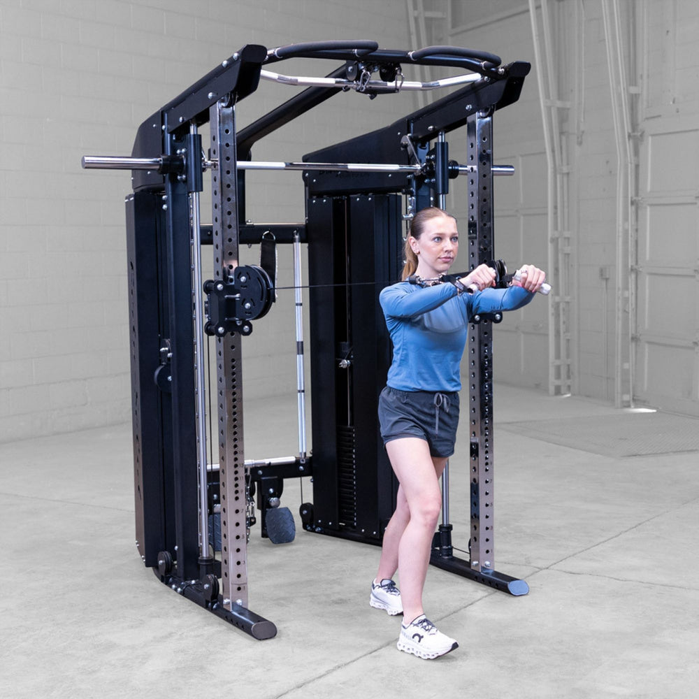 A woman in athletic wear is using a black and silver Body-Solid GSFR100 multi-functional home gym cable machine with a pull-up bar, set in a concrete-floored warehouse gym.