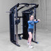 A woman in athletic wear is using a black and silver Body-Solid GSFR100 multi-functional home gym cable machine with a pull-up bar, set in a concrete-floored warehouse gym.
