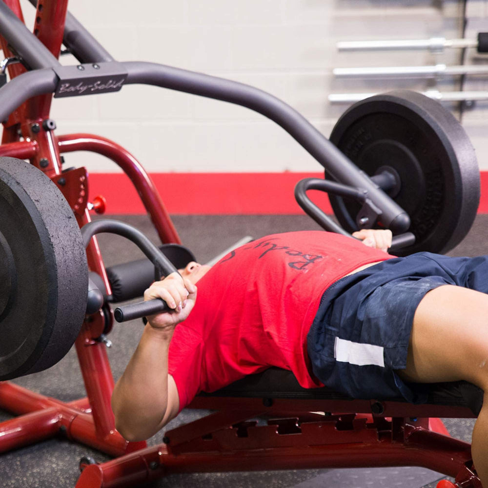 Body-Solid Corner Leverage Gym GLGS100B Man lying on a bench performing a barbell bench press using a gym machine with black weight plates.

