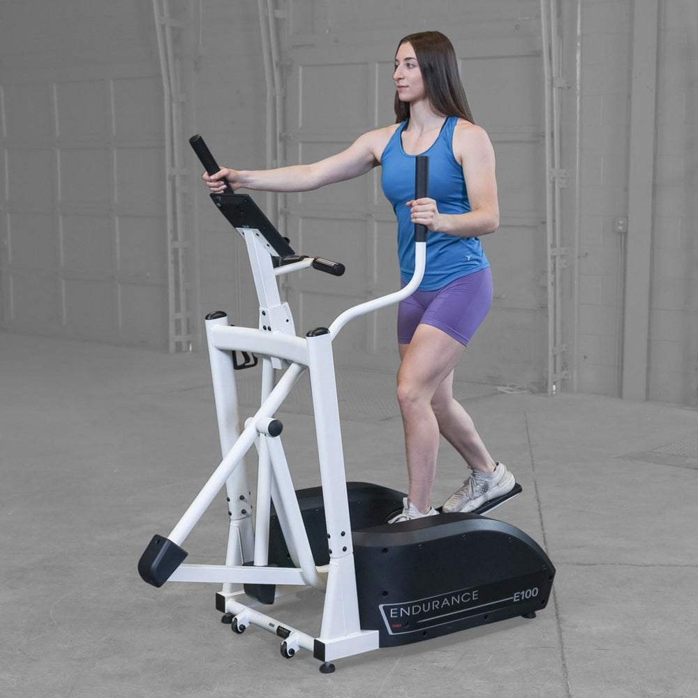 Body-Solid Endurance Center Drive Elliptical E100 Front-angle view of woman performing workout on Endurance E100 elliptical trainer inside light-gray gym environment.
