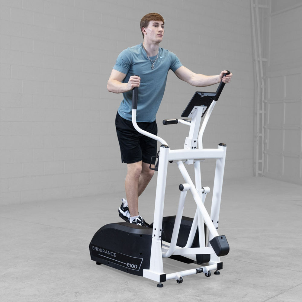 Body-Solid Endurance Center Drive Elliptical E100 Man exercising on Endurance E100 elliptical trainer, demonstrating full stride motion in industrial gym space.
