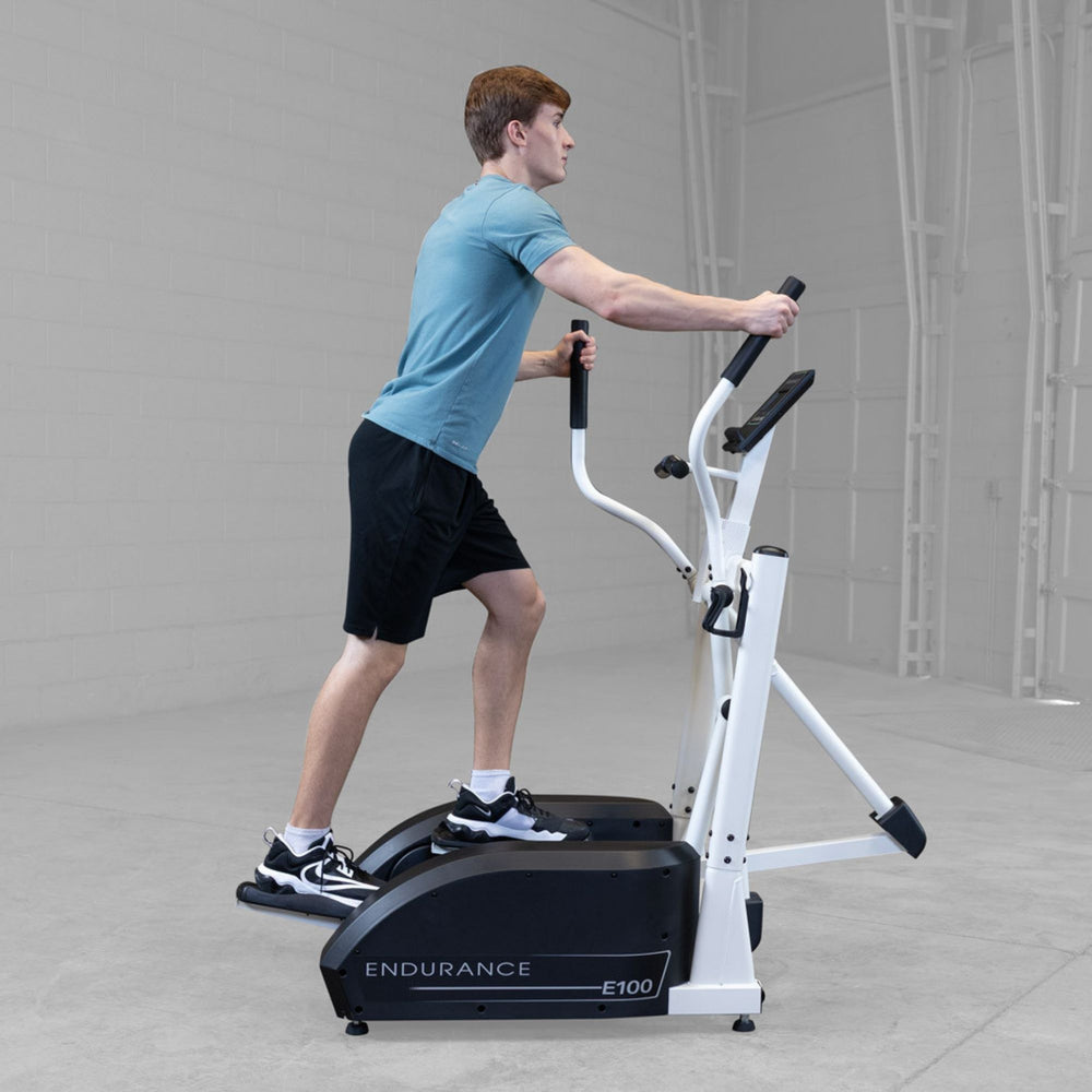 Body-Solid Endurance Center Drive Elliptical E100 Man mid-stride on Endurance E100 elliptical trainer during cardio workout inside a modern fitness setting.
