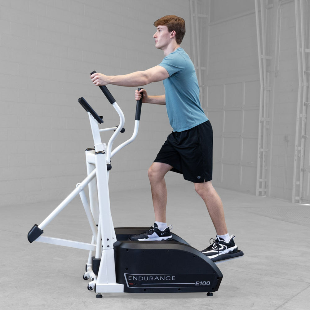 Body-Solid Endurance Center Drive Elliptical E100 Man using Endurance E100 elliptical trainer with hands on moving handles during cardio workout session.
