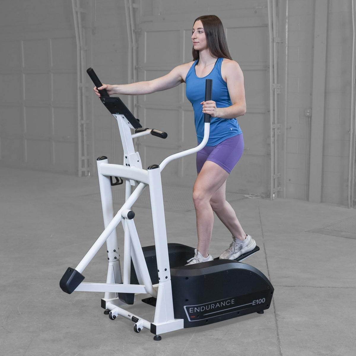 Body-Solid Endurance Center Drive Elliptical E100 Woman using Endurance E100 elliptical trainer, side view showing stride and full upper-body engagement.

