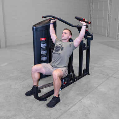 Body-Solid PCL2 Multi Press S2MPX Man performing seated on a shoulder press machine pushing handles upward in a gym setting
