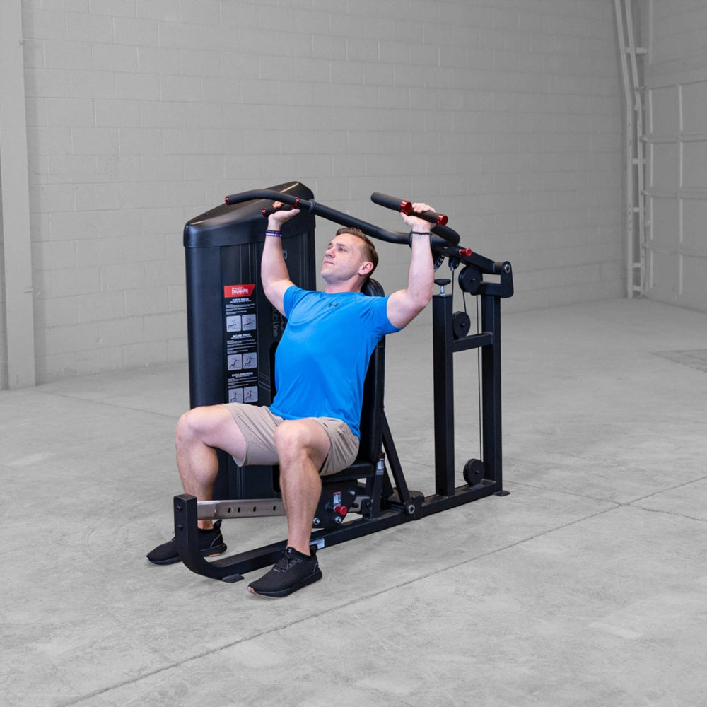 Body-Solid PCL2 Multi Press S2MPX Man seated on a shoulder press machine pushing handles upward in a gym setting
