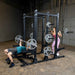Body-Solid Pro ClubLine Double Half Rack SPR500DBL Man bench pressing and woman squatting using Body-Solid SPR500DBL double training rack equipped with weight plates and barbell supports.