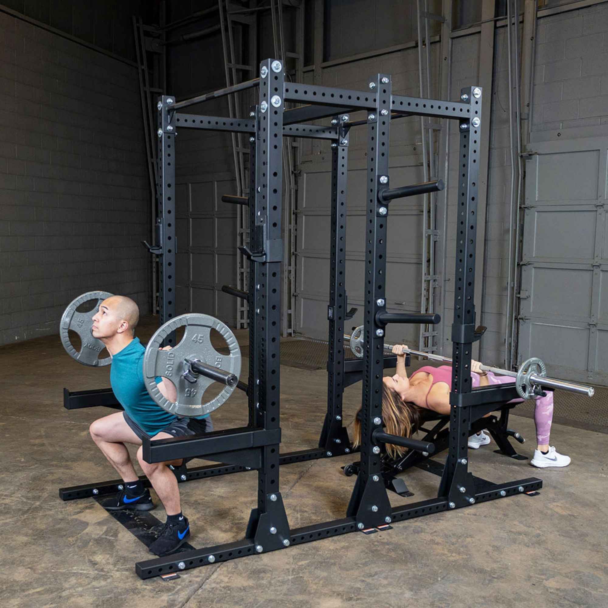 Body-Solid Pro ClubLine Double Half Rack SPR500DBL Man performing barbell squat and woman bench pressing on Body-Solid SPR500DBL power rack in industrial gym environment.
