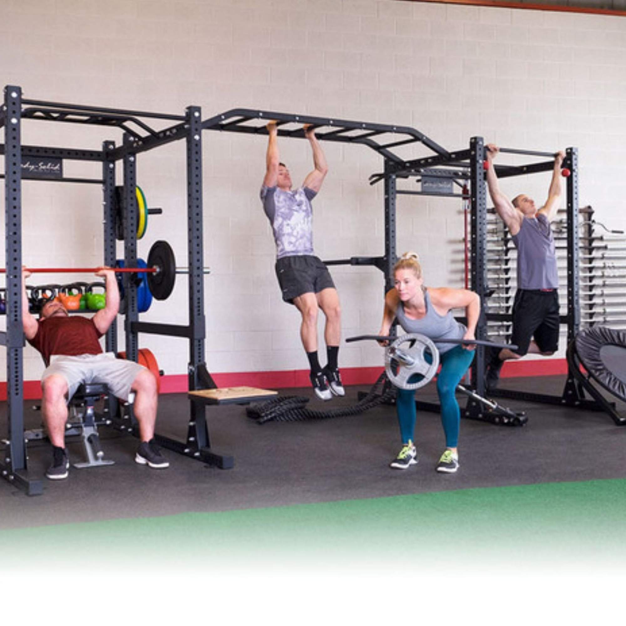 Body-Solid Pro ClubLine Double Power Racks with Rear Extensions and Monkey Bars with Safety Straps SPR1000SSDBBACK A group of four people exercising on a large, multi-station black fitness rig in a gym. On the left, a man is bench pressing. 