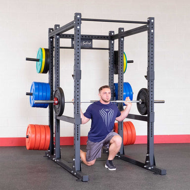 Body-Solid Pro ClubLine SPRBACK Power Rack Rear Extension SPRBACK Man performing a barbell squat inside a Body-Solid power rack loaded with colorful weight plates in a gym setting.