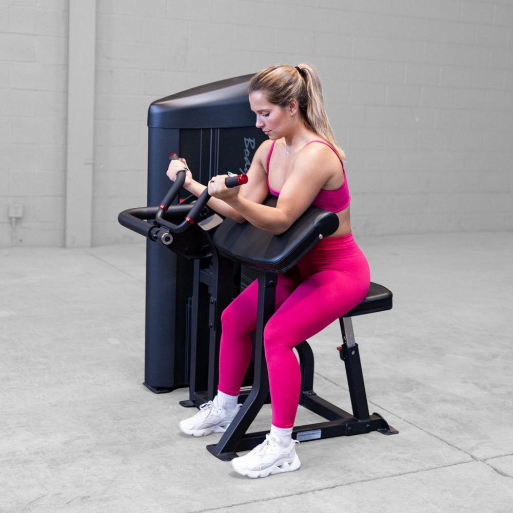 Body-Solid Pro ClubLine Series 2 Arm Curl S2ACX Woman performing seated preacher curls on a Body-Solid ProClubLine arm curl machine while wearing a bright pink workout outfit.