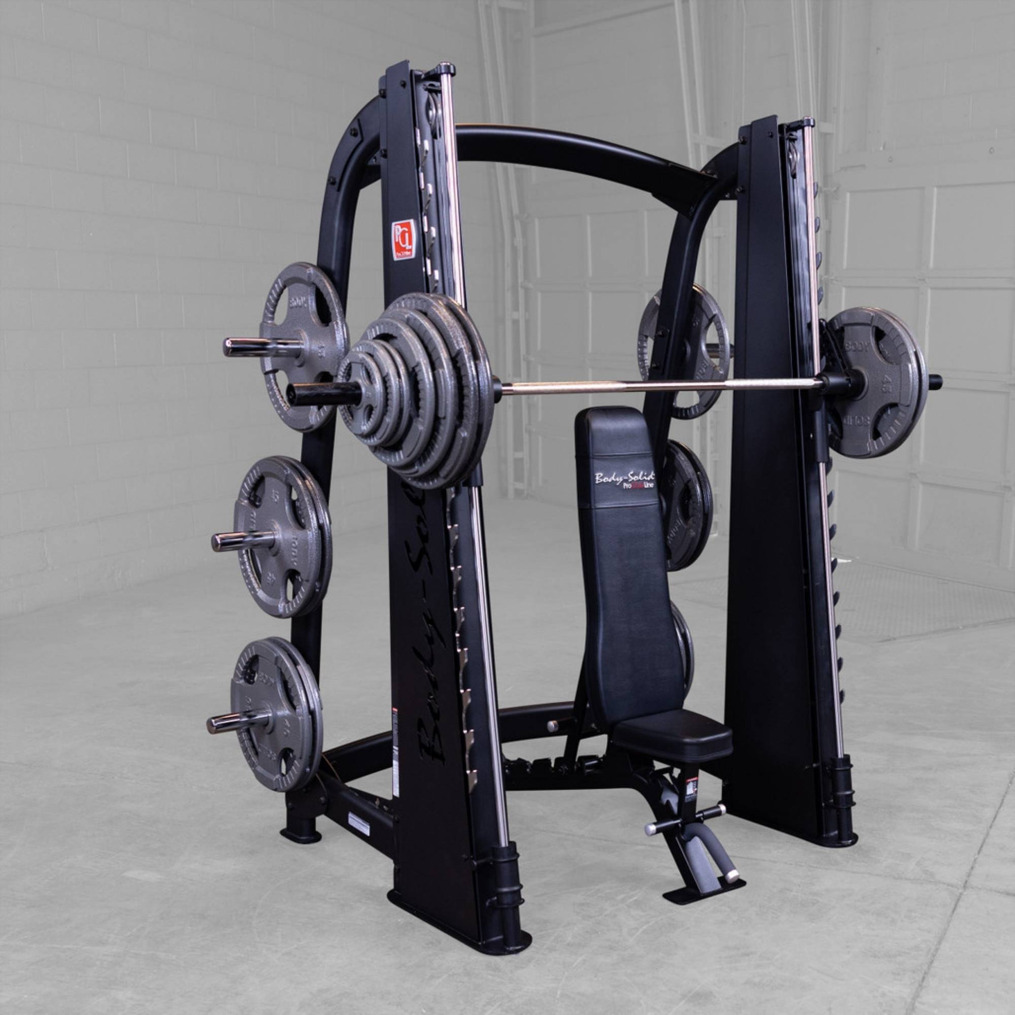 Body-Solid Proclub Counter Balanced Smith Machine SCB1000B A Smith machine loaded with multiple weight plates on storage pegs, bench placed inside the rack in a gym.
