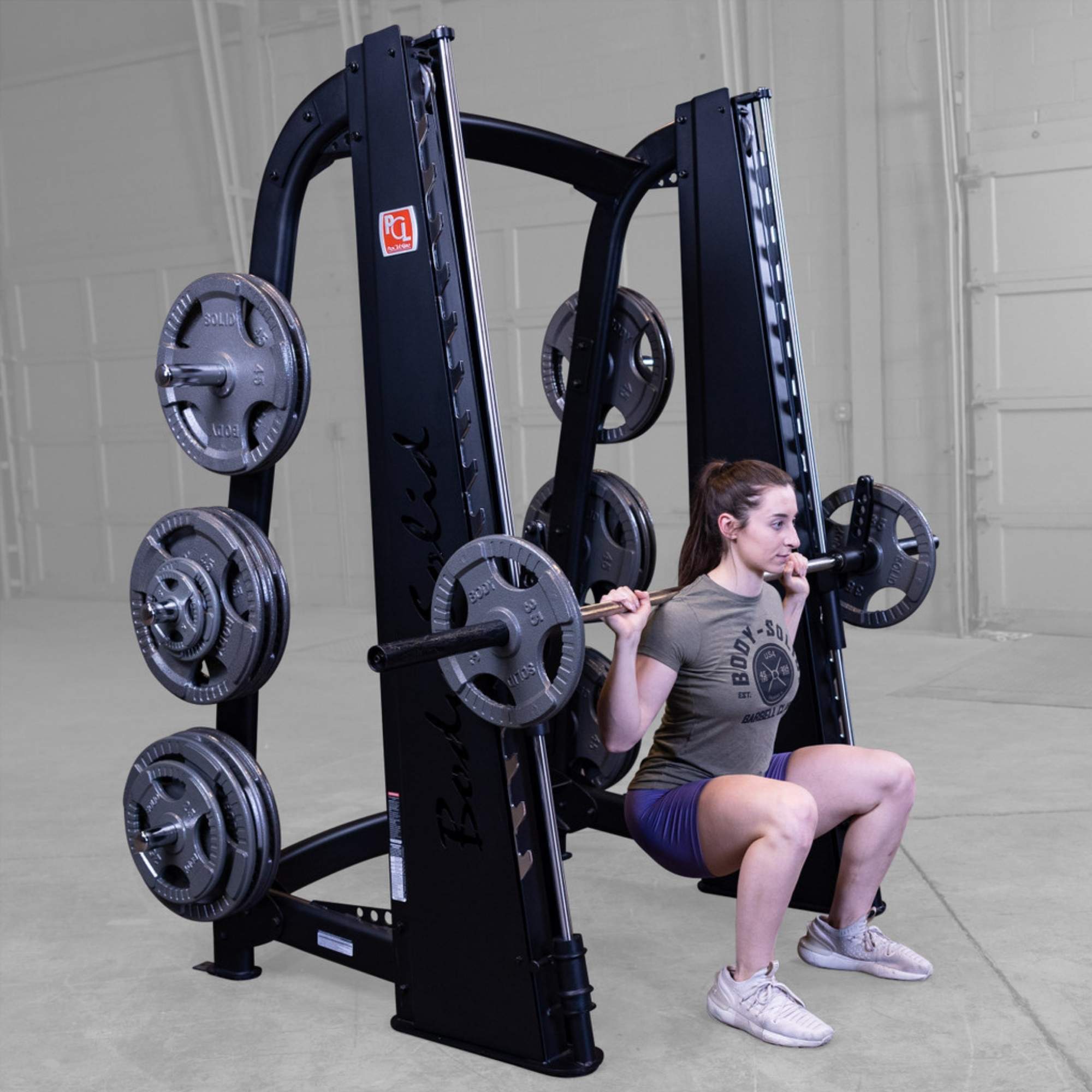 Body-Solid Proclub Counter Balanced Smith Machine SCB1000B A woman doing a Smith machine back squat with the bar positioned on her shoulders, squatting low in a gym.
