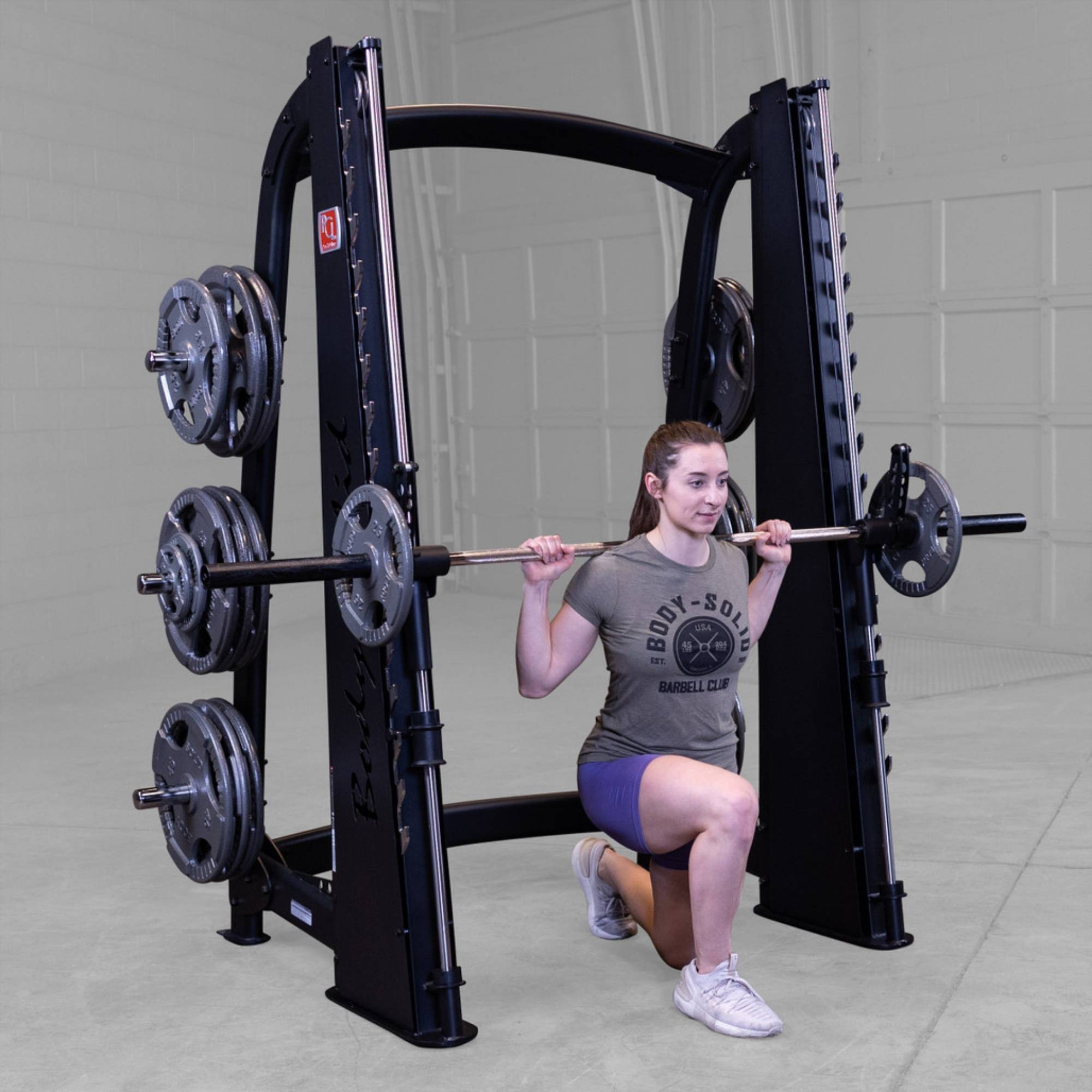 Body-Solid Proclub Counter Balanced Smith Machine SCB1000B A woman doing a barbell walking lunge on a Smith machine, holding the bar across her shoulders in a gym.
