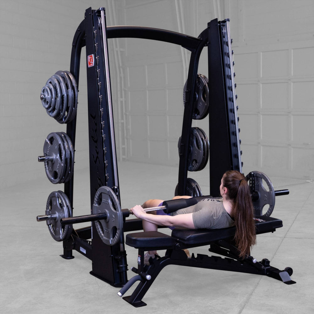 Body-Solid Proclub Counter Balanced Smith Machine SCB1000B A woman lying on a bench performing a barbell hip thrust inside a Smith machine in a gym.
