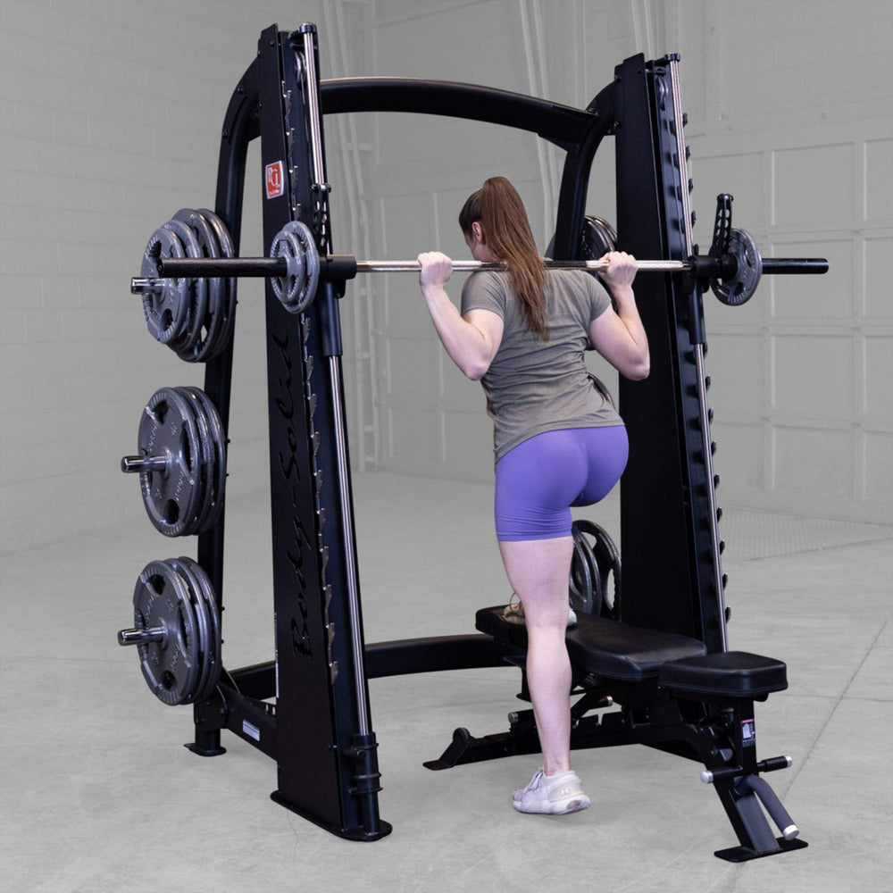 Body-Solid Proclub Counter Balanced Smith Machine SCB1000B A woman standing behind a bench, positioning a barbell across her shoulders on a Smith machine, preparing for step-ups.

