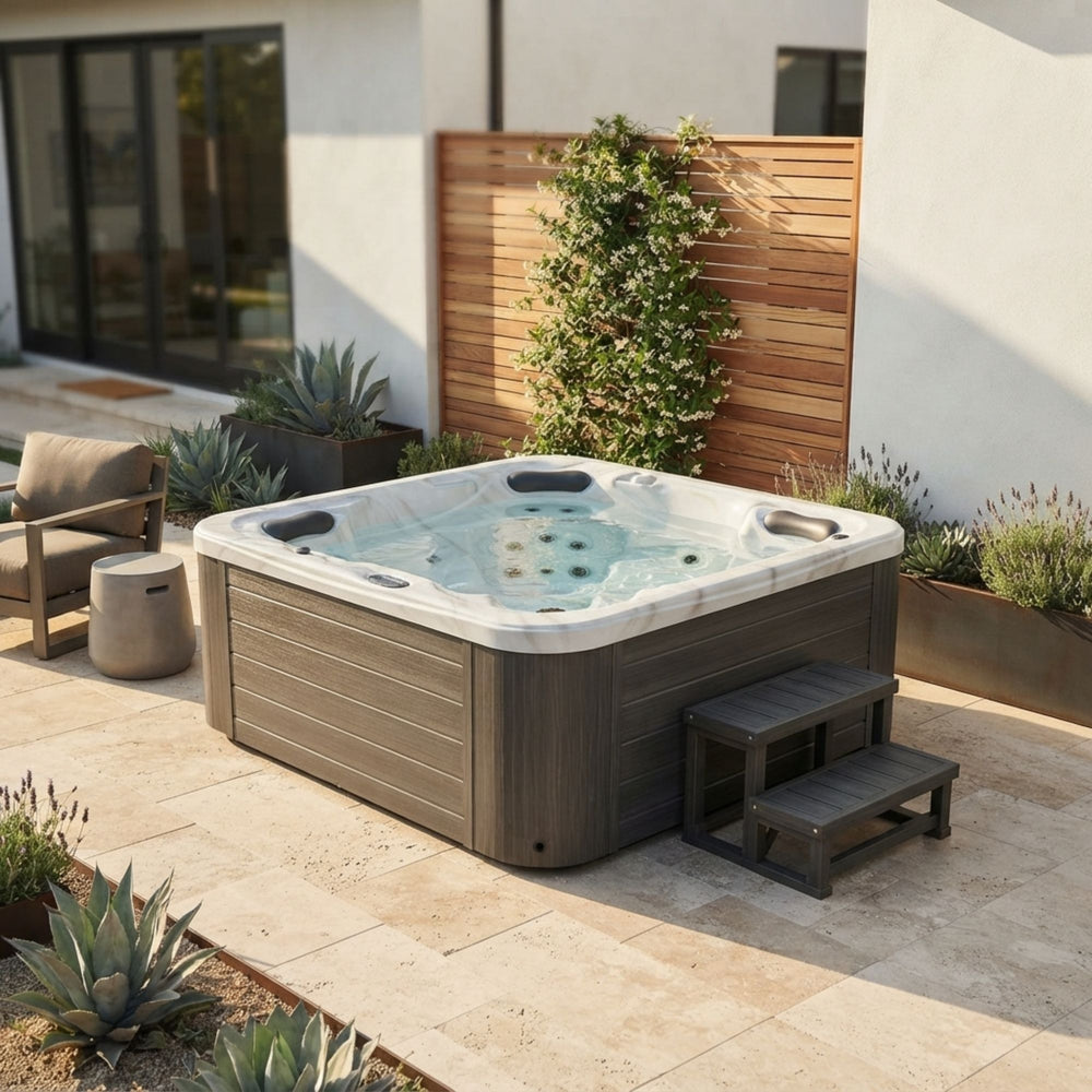 A Luxury Spa Estes outdoor hot tub with a gray wood-look exterior and white interior, set on a stone patio beside a modern house. The tub has dark gray entry steps and is surrounded by plants and a wooden fence.