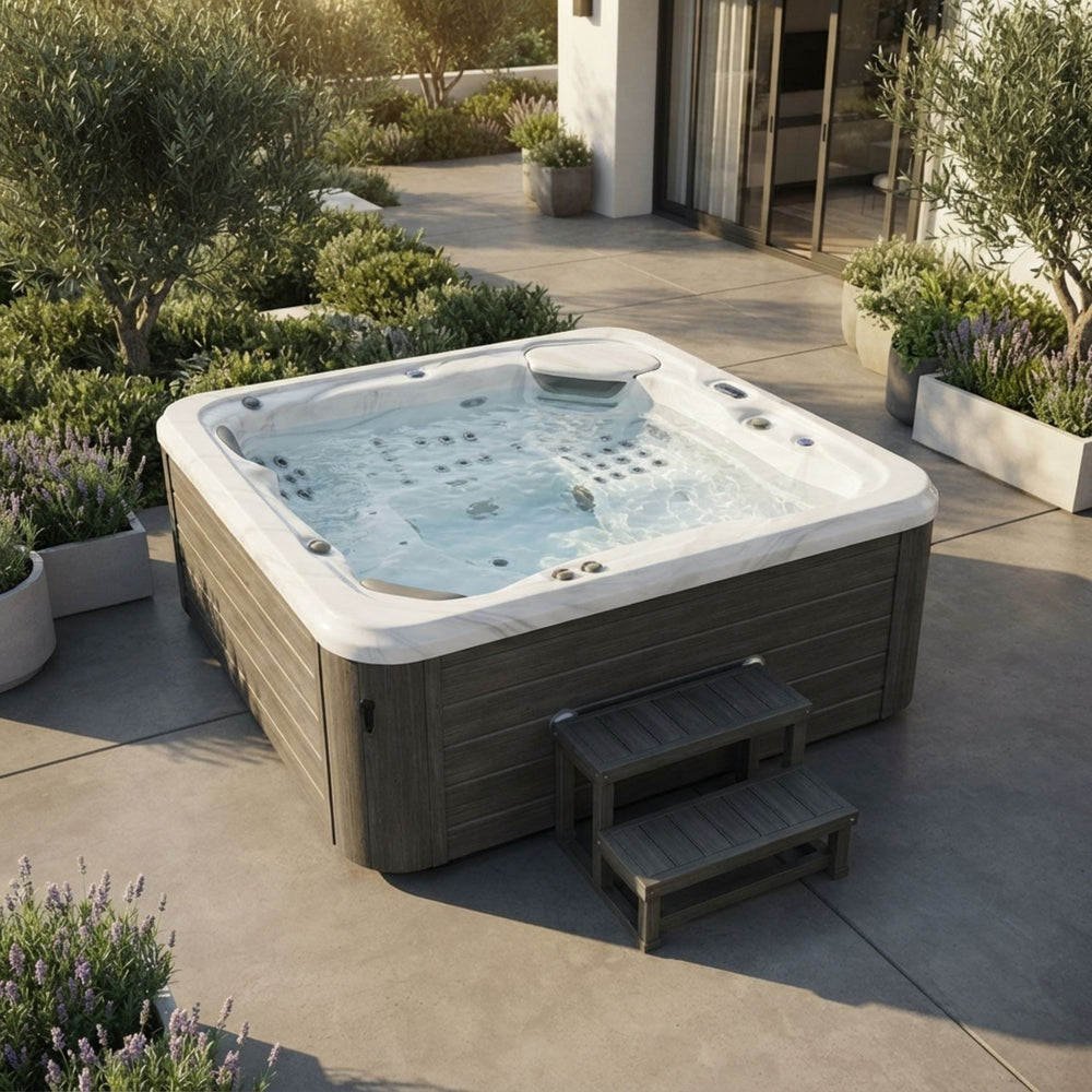 A Luxury Spa Infinity 5-person outdoor hot tub with gray wood-look siding and a white top rim, shown on a patio in a landscaped backyard. The tub is filled with bubbling water, and two matching gray steps are placed beside it