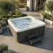 A Luxury Spa Infinity 5-person outdoor hot tub with gray wood-look siding and a white top rim, shown on a patio in a landscaped backyard. The tub is filled with bubbling water, and two matching gray steps are placed beside it