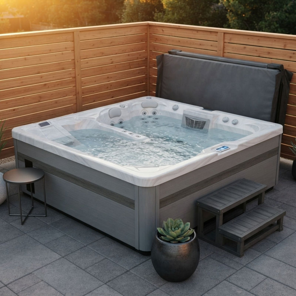 A rectangular outdoor Luxury Spas USA Marquis Hot & Cold Contrast Therapy Spa hot tub with bubbling water, set on a paved patio corner with a wooden fence, a small side table, two wooden steps, and a potted succulent. The hot tub has a gray exterior and white interior, with a dark gray cover folded behind it.