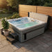 A rectangular outdoor Luxury Spas USA Marquis Hot & Cold Contrast Therapy Spa hot tub with bubbling water, set on a paved patio corner with a wooden fence, a small side table, two wooden steps, and a potted succulent. The hot tub has a gray exterior and white interior, with a dark gray cover folded behind it.