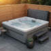A rectangular outdoor Luxury Spas USA Marquis Hot & Cold Contrast Therapy Spa hot tub with bubbling water, set on a paved patio corner with a wooden fence, a small side table, two wooden steps, and a potted succulent. The hot tub has a gray exterior and white interior, with a dark gray cover folded behind it.