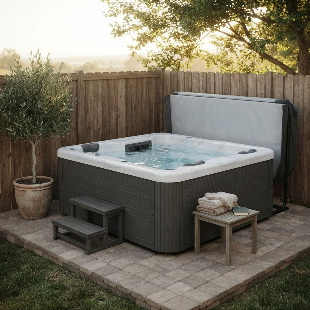 A Luxury Spas USA Allure 3-Person outdoor hot tub with a white interior and jets, set on a paved patio in a backyard with a wooden fence, a small bench, a side table with folded towels, and a potted tree.
