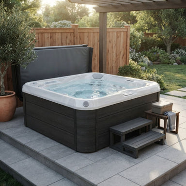 A dark gray outdoor Luxury Spas Liberty 5-Person hot tub with a white interior and bubbling water, set on a tiled patio in a landscaped backyard with a wooden fence, plants, and a small wooden step stool.