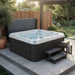 A dark gray outdoor Luxury Spas Liberty 5-Person hot tub with a white interior and bubbling water, set on a tiled patio in a landscaped backyard with a wooden fence, plants, and a small wooden step stool.