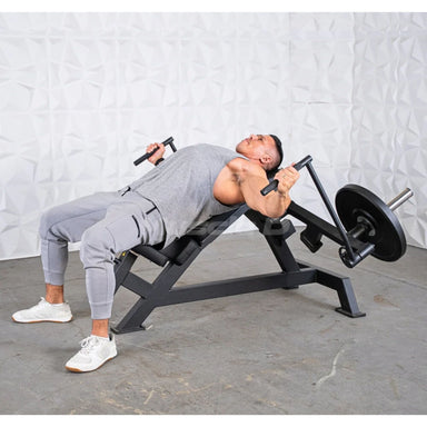 A man in a grey athletic outfit performing a weight leverage pec fly bench press on a black adjustable weight bench in a gym setting with a textured white wall and concrete floor.